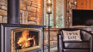 Une pièce chaleureuse avec un mur en pierre, une cheminée où l’on peut voir les flammes, un fauteuil en cuir avec un coussin et un téléviseur.