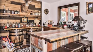 A rustic bar area with wooden shelves displaying bottles, a deer head mounted on the wall, and a large window. - Stepaway Vacation Rentals- Happy Hours Pet Friendly (Eagle River)