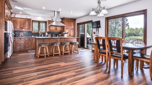 A spacious kitchen with wooden cabinets, a large island with stools, a dining area with a table and chairs, and a view through large windows. - Stepaway Vacation Rentals- Happy Hours Pet Friendly (Eagle River)