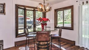 A dining area with a round glass table, four chairs, a chandelier, and large windows with curtains. - Stepaway Vacation Rentals- Happy Hours Pet Friendly (Eagle River)