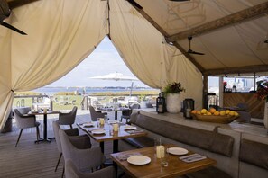 Ein Essbereich im Freien mit Tischen und Stühlen, Blick auf das Wasser und eine Holzterrasse.