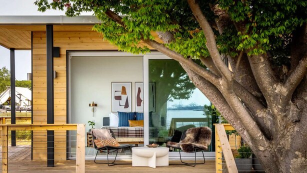 Una casa moderna con terraza de madera, puerta de cristal que da a un espacio de estar con sofá y mesa, y un árbol grande al frente.