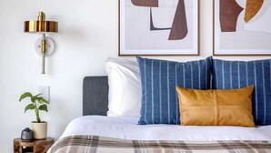 Una cama bien hecha con una manta escocesa, una planta en la mesita de noche y una lámpara de pared.