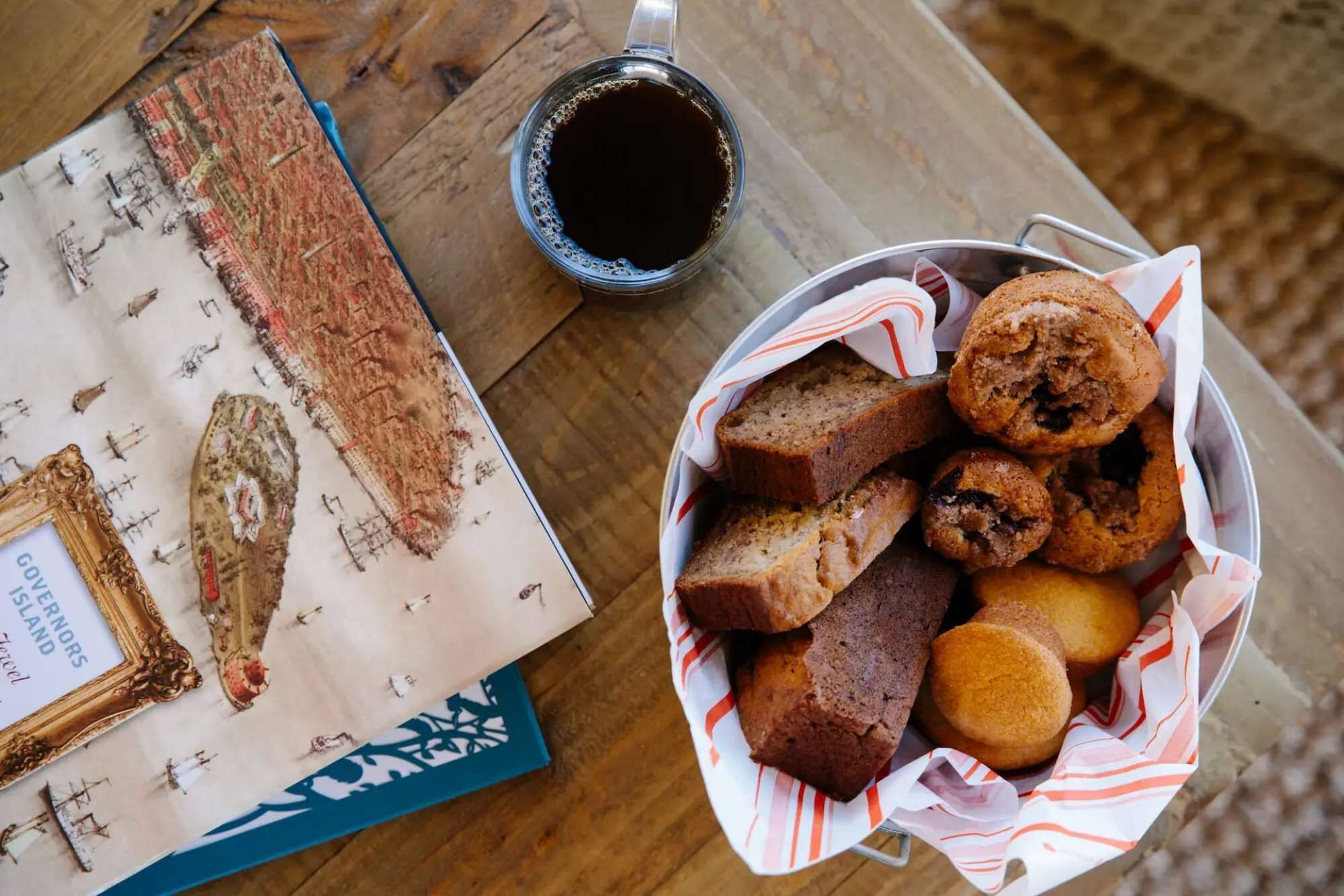 Ein Tisch mit einer Korb voller Muffins und einer Tasse Kaffee, eine gerahmte Karte und ein Buch mit dem Titel 'GOVERNORS ISLAND'.
