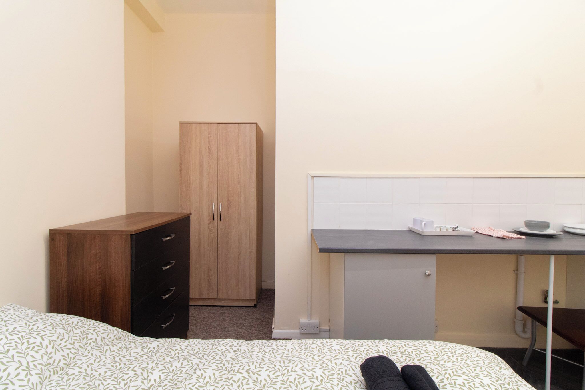 Una habitación de hotel pequeña con una cama, un armario de madera, una kitchenette con mostrador y una pared de azulejos blancos.