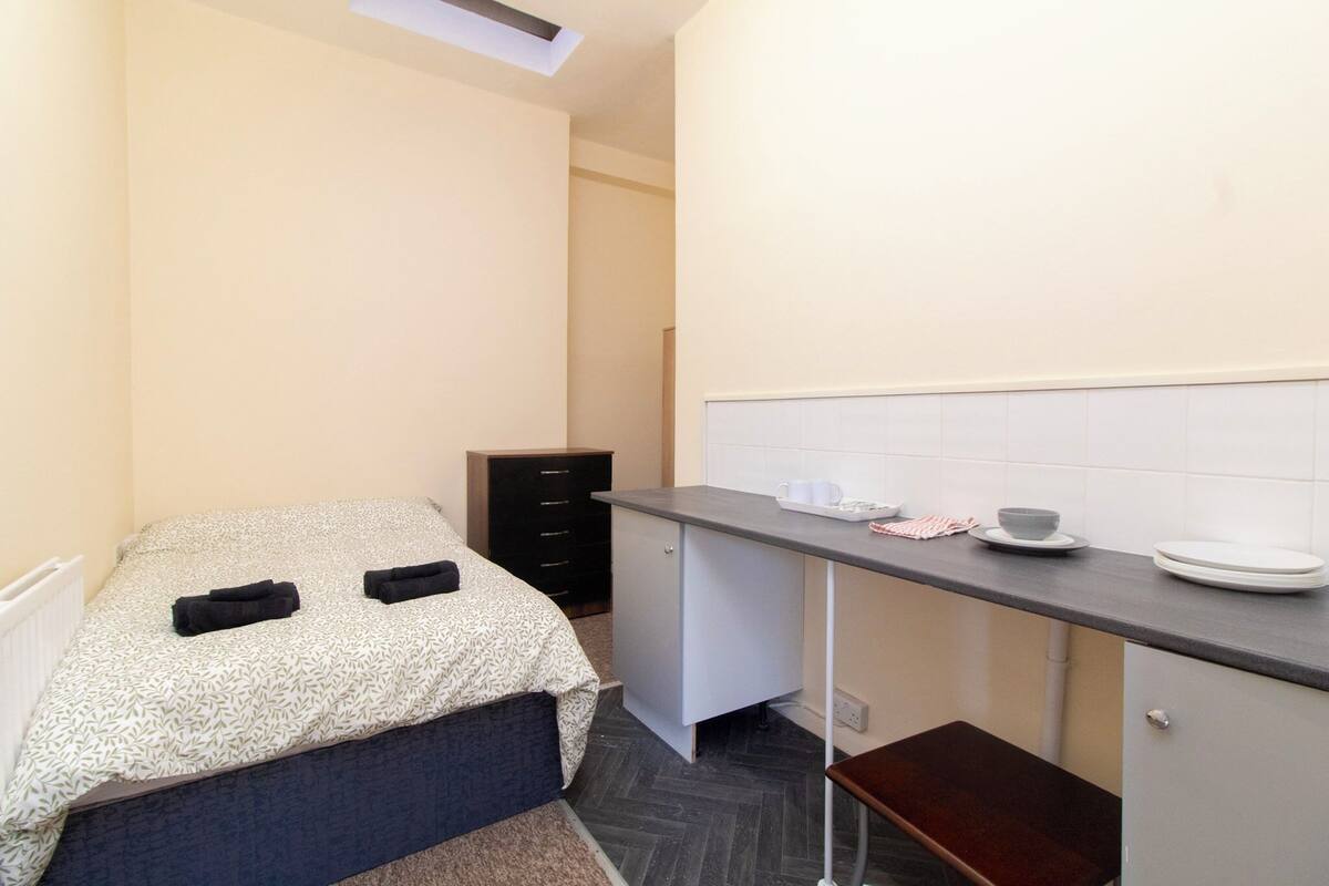 Een kleine, praktische kamer met een bed, bureau en kitchenette.