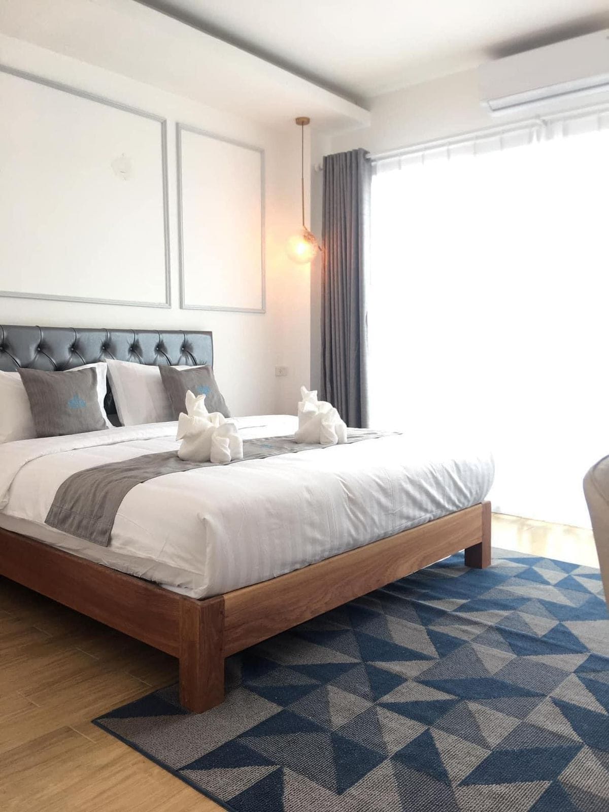 A neatly made bed with a wooden frame, a geometric patterned rug, and a large window with curtains.
