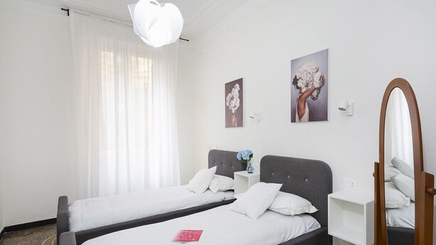 Habitación de hotel con dos camas individuales, una mesita de noche con un florero, un espejo grande y dos cuadros colgados en la pared.