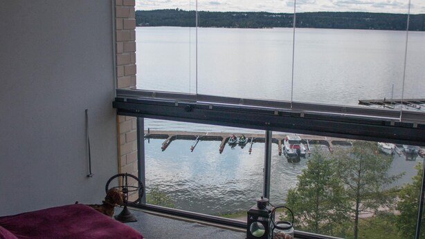 A room with a large window overlooking a body of water, a wooden table with a purple cloth, and a floral rug. - Kotimaailma Deluxe Apartment Lake View (Lahti)