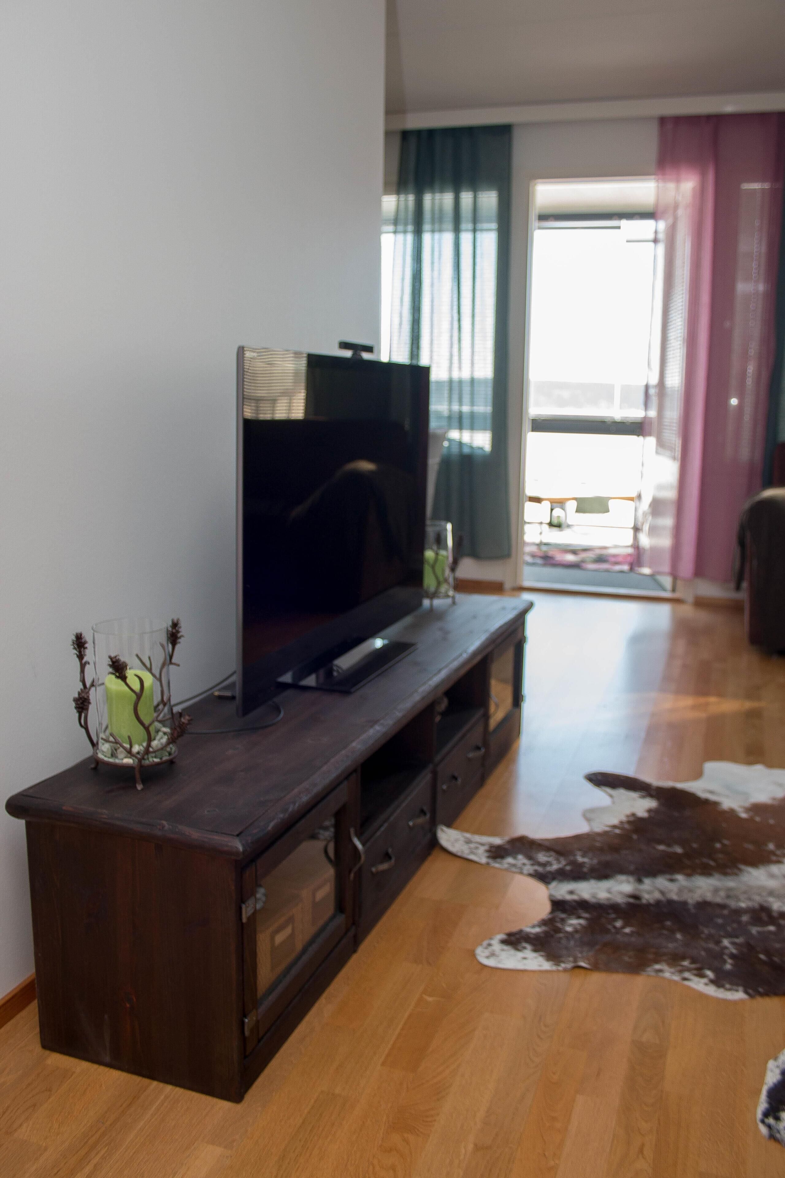 Una sala de estar con un mueble de entretenimiento de madera oscura, un televisor de pantalla plana, un florero de vidrio con plantas secas, un tapete de cuero de vaca y una puerta corrediza de cristal que da a un balcón.