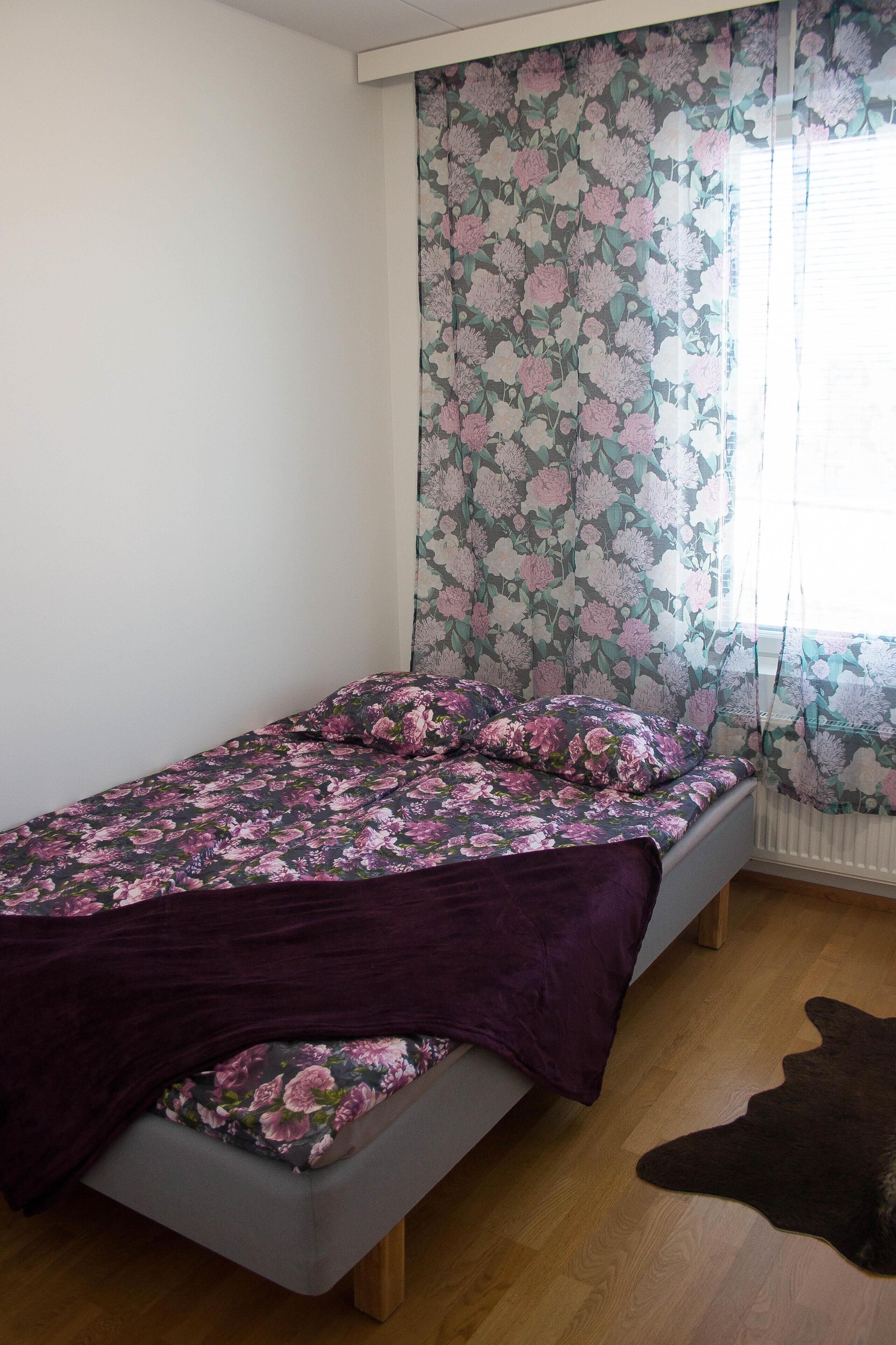 A single bed with floral bedding, a window with floral curtains, and a wooden floor.