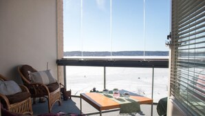 A room with a view of a snowy landscape through large windows. There is a wooden table with glassware and a chair with cushions. - Kotimaailma Deluxe Apartment Lake View (Lahti)