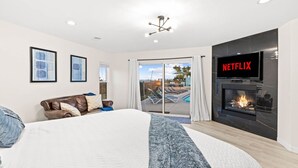 Une chambre moderne avec un grand lit, une cheminée, un téléviseur affichant « NETFLIX », et une vue sur une piscine et un espace extérieur.