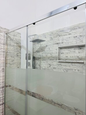 Une douche fermée par des parois de verre, avec un mur imitant le marbre et une étagère intégrée.