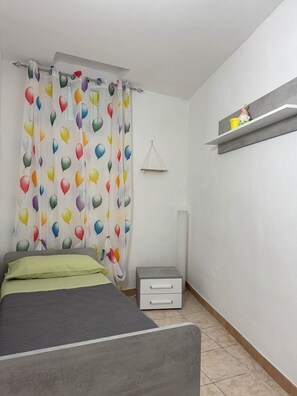 Une chambre à coucher comprenant un lit, une table de chevet, une étagère fixée au mur et un rideau orné d’un motif représentant des ballons.
