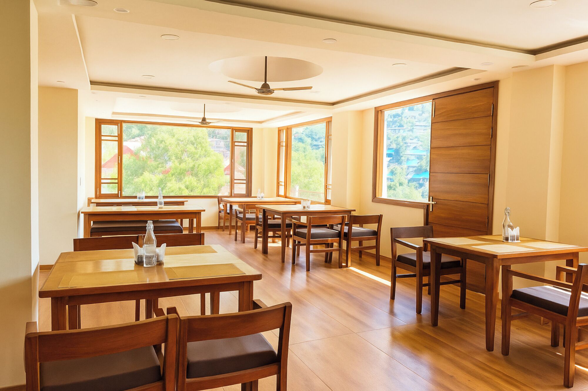 A spacious dining area with wooden tables and chairs, large windows offering a view of greenery, and a ceiling fan.