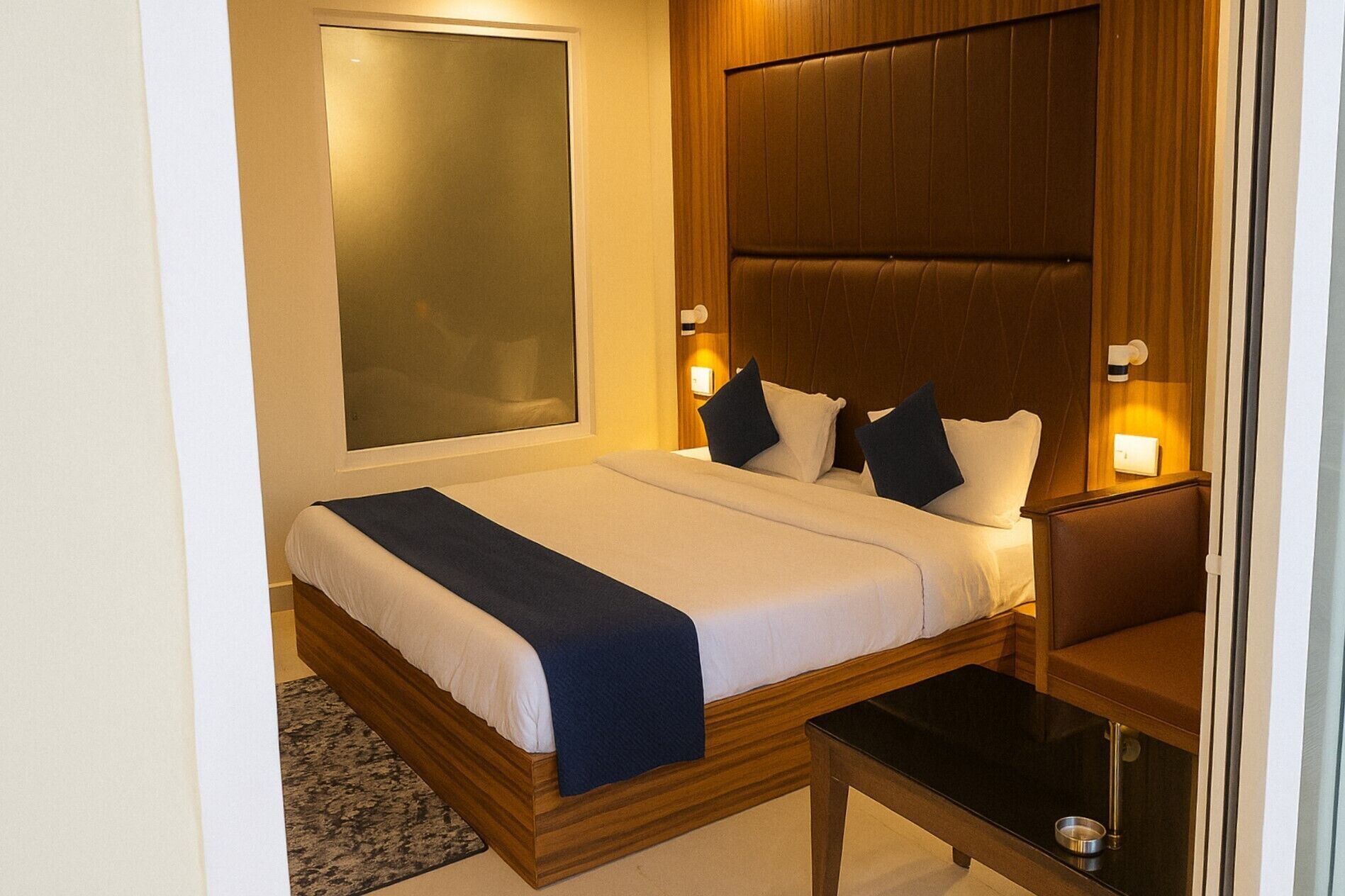 Cama bem arrumada com cabeceira de madeira, duas luminárias laterais e uma mesinha com uma tigela.