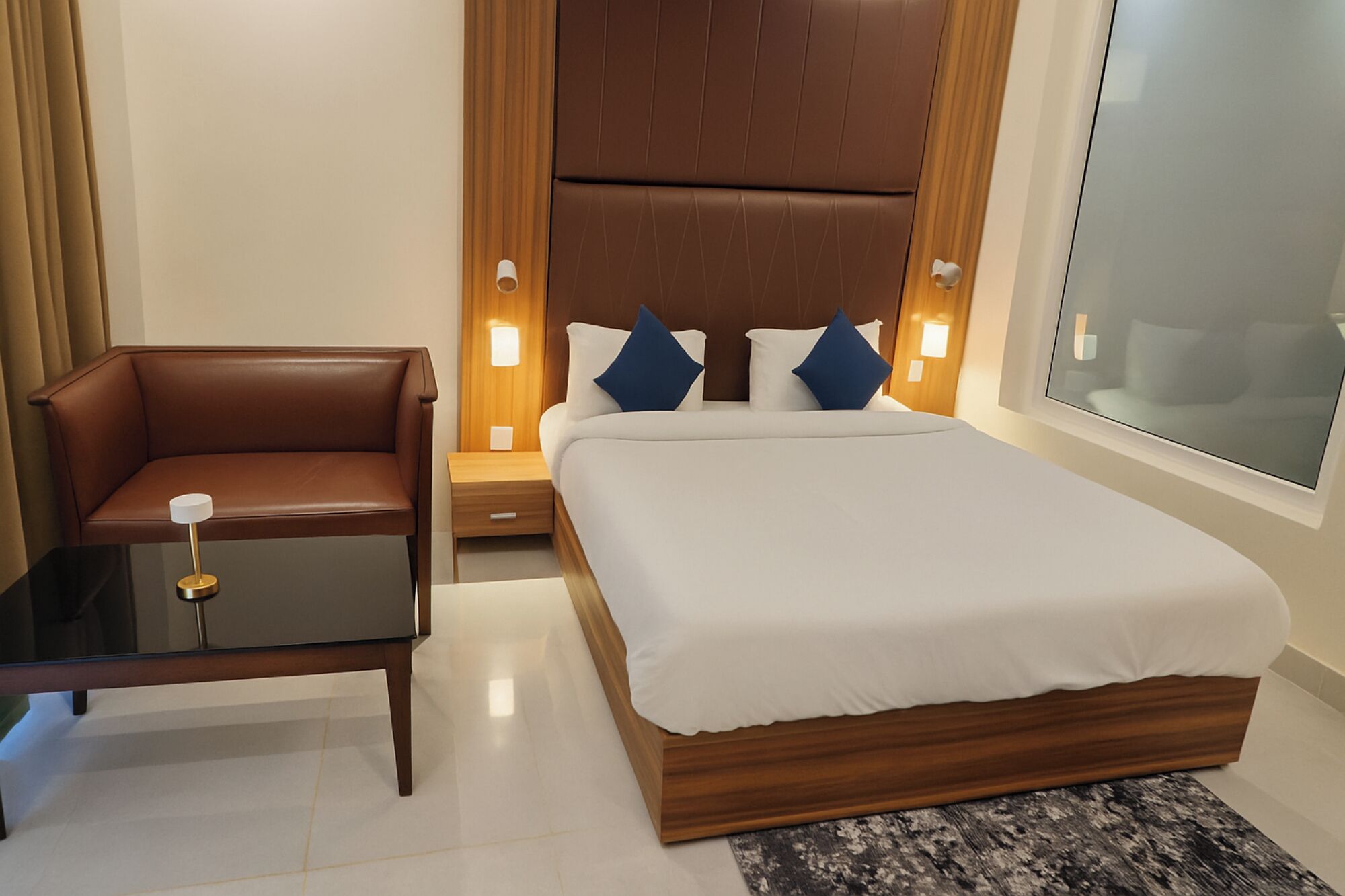 A modern hotel room with a leather chair, a bedside table, a wooden headboard, and a large bed with white bedding.