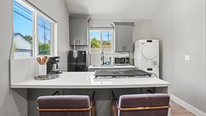 A modern kitchen with a large island, grey cabinets, a black refrigerator, a white microwave, and a white dishwasher. There are two bar stools with a purple upholstery and a window providing a view of the outside.