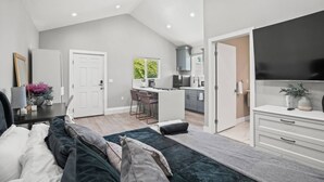 A modern bedroom with a large bed, a TV, and a kitchenette in the background.
