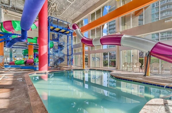 An indoor swimming pool with a water slide, a children's play area, and a view of a building with balconies.