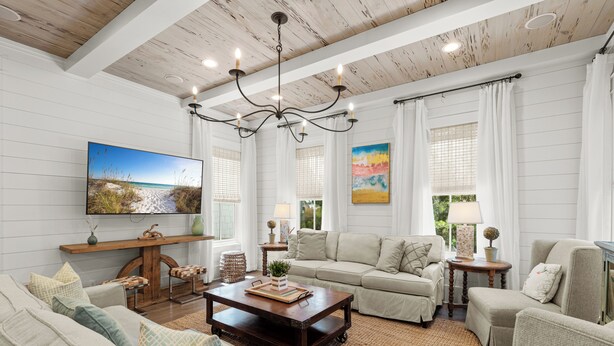 A spacious living room with a high ceiling, a large chandelier, and a television mounted on the wall. There are multiple seating areas, including a sofa and chairs, and a coffee table in the center. - Welcome to Azalea Abbey! Private Pool, Golf Cart, Walk to Beach, Seaside (Santa Rosa Beach)