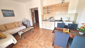 Una sala de estar con un sofá, una mesa de centro y un comedor con sillas y una mesa. Hay una zona de cocina con armarios y un lavamanos.