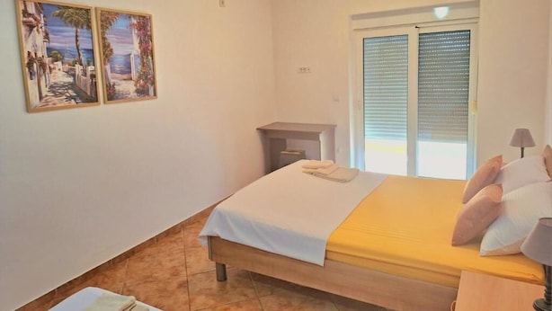 Un dormitorio con una cama, una mesita de noche, un ventanal con persianas y dos cuadros colgados en la pared.