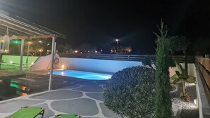 A nighttime view of a swimming pool area with a covered seating area and a view of the city skyline in the background. - Boho Sisi Haven- Poolside Oasis Garden Retreat (Agios Nikolaos)