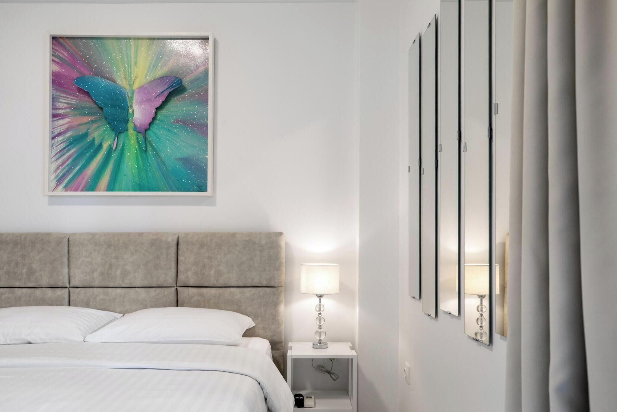 Quarto de hotel com cama, cabeceira, mesa de cabeceira com abajur, espelho fixado na parede e uma grande pintura abstrata.