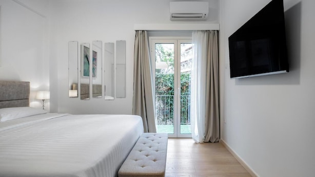 A modern hotel room with a large bed, a flat-screen TV mounted on the wall, a bench, and a sliding glass door leading to an outdoor area. - Acropolis Museum Apartment by A&A Residences (Athens)