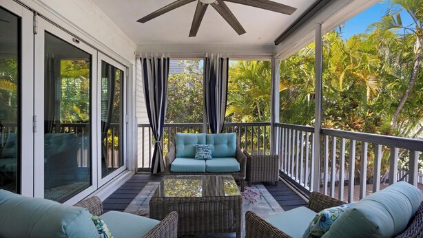 A covered porch with a ceiling fan, a light blue sofa, wicker furniture, and a view of tropical foliage.