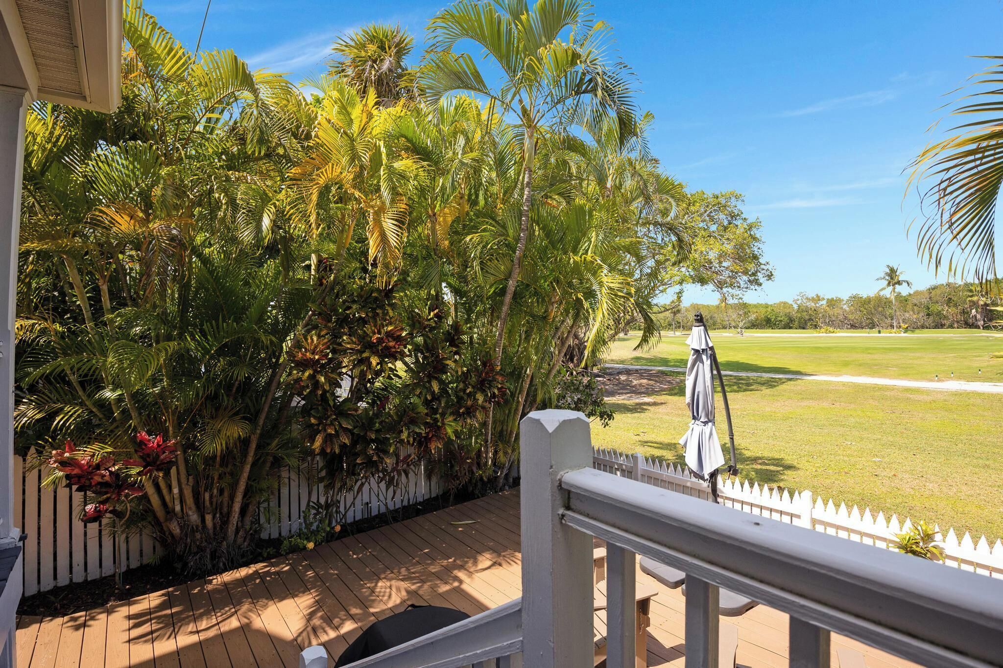 A wooden deck with a white picket fence, lush tropical plants, and a golf course in the background.