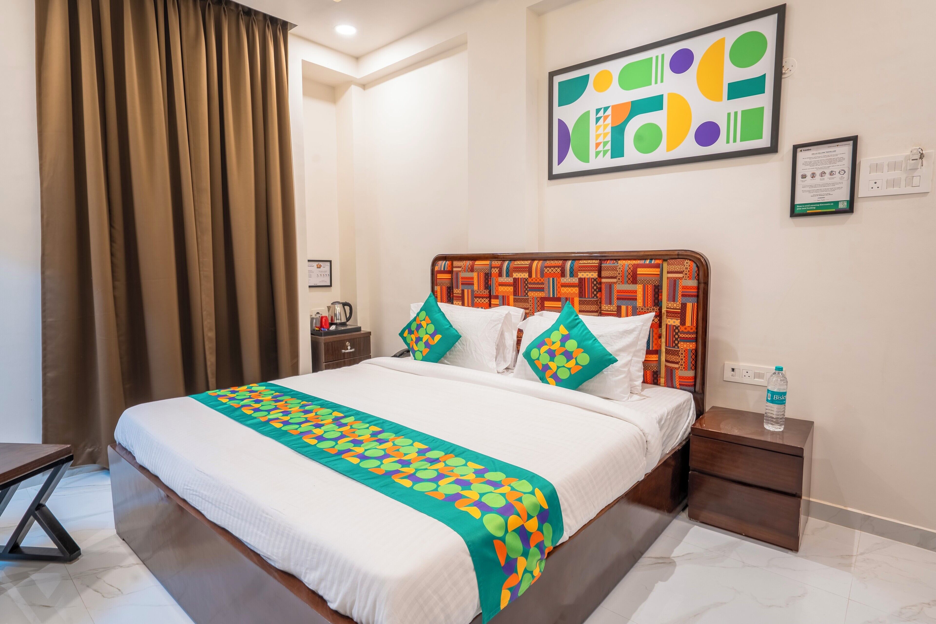 A hotel room with a large bed, a wooden headboard, a colorful bedspread, a nightstand with a water bottle, and a painting on the wall.