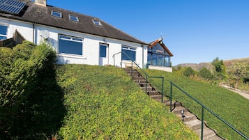 Situated on a hillside, this two-story house features a sloped roof and solar panels, surrounded by a well-maintained garden and a stone staircase leading to the entrance.