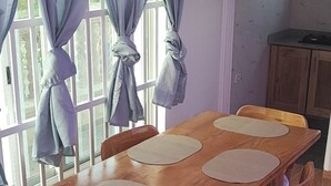 Ein Essbereich mit Holztisch und Stühlen, einem Fenster mit Vorhängen sowie einer Küche im Hintergrund.