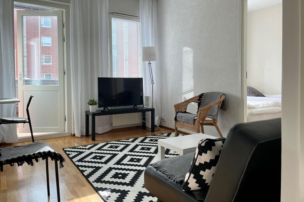A modern living room with a flat-screen TV, a wicker chair, a sofa with a patterned throw, and a patterned rug.