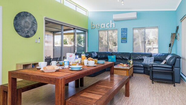 A modern living room with a wooden dining table, a black leather sofa, and a large window. - Break-A-Way COASTAL COMFORT IN LANCELIN (Lancelin)