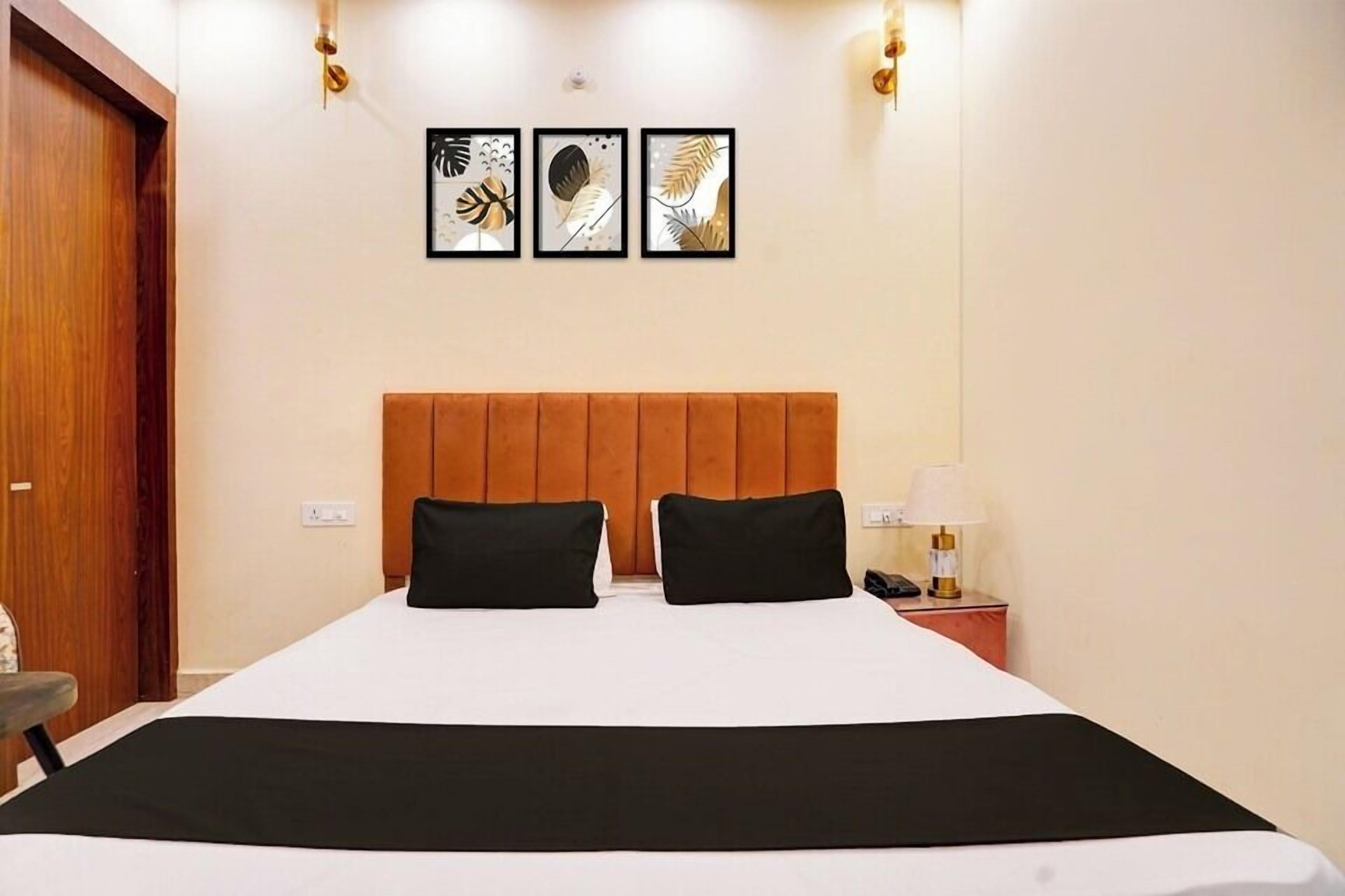 A hotel room with a bed, two black pillows, a wooden headboard, a nightstand with a lamp, and three framed leaf artworks on the wall.