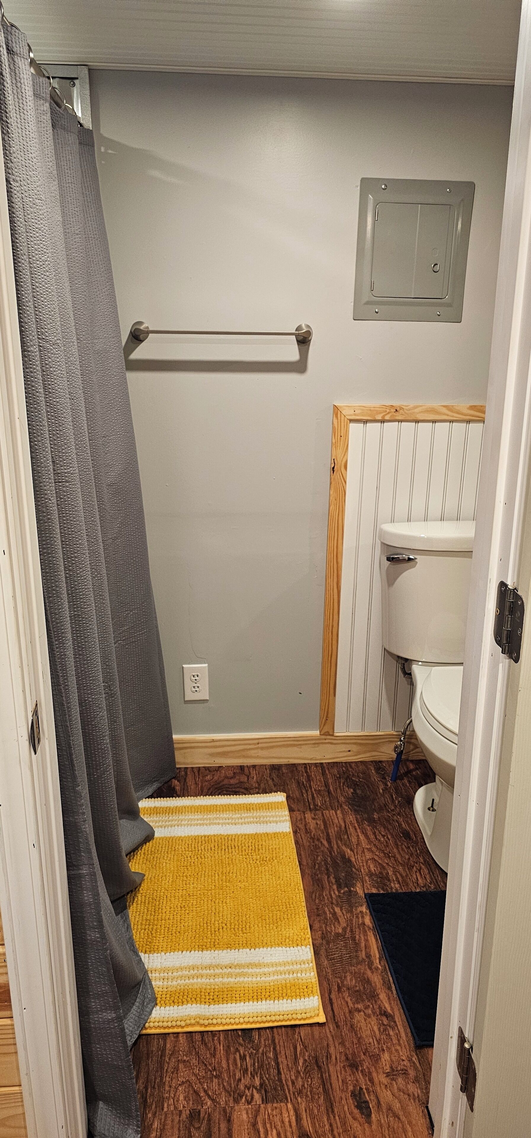 Een kleine badkamer met een houten vloer, een geel-wit gestreept tapijt, een wit toilet en een grijze douchegordijn.