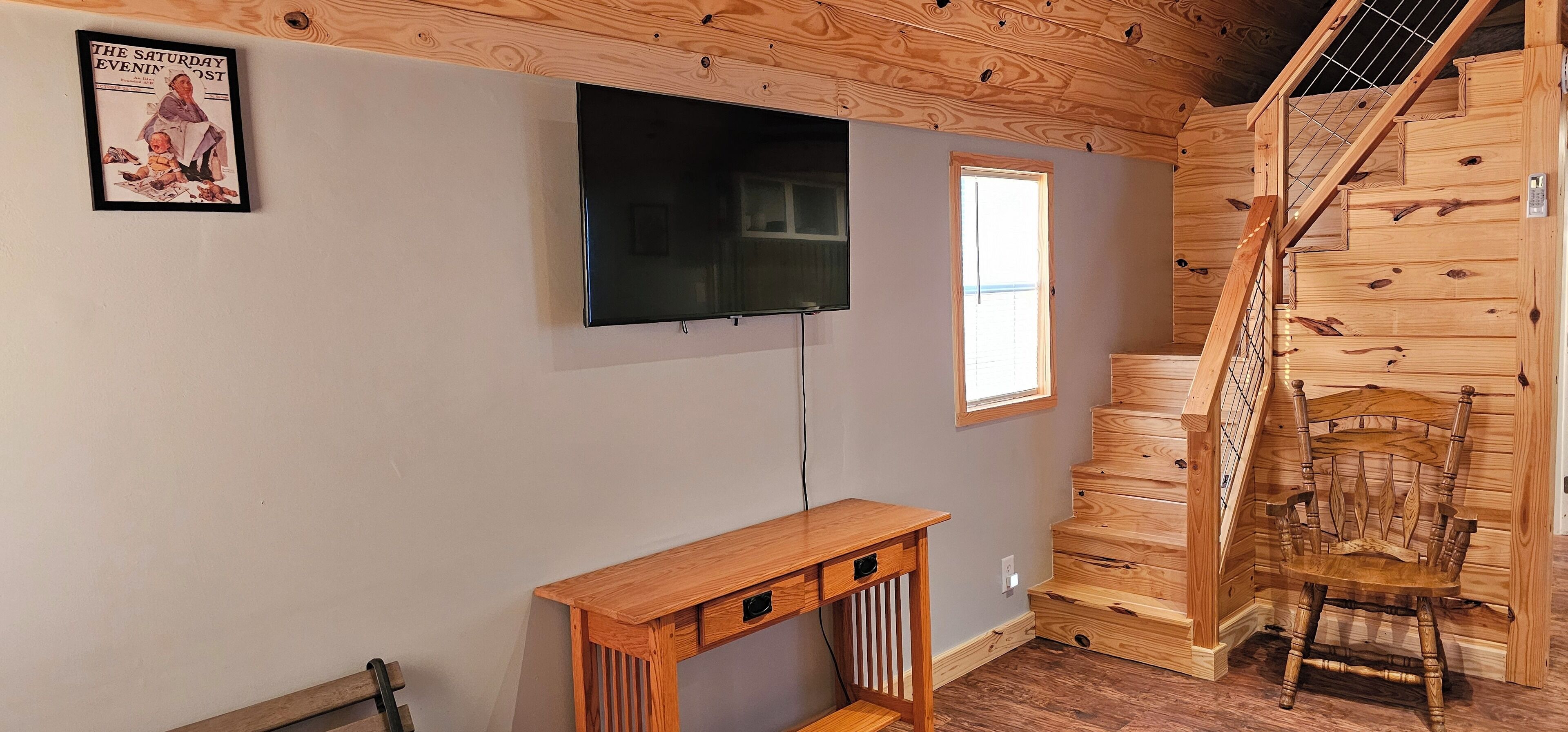 A room with a wooden staircase, a TV mounted on the wall, and a small wooden table.