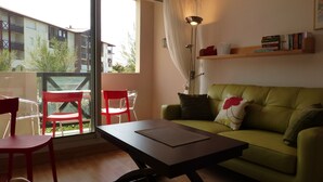 Una sala de estar con un sofá verde, una mesa de centro de madera y una silla roja. Se ve una ventana con vistas a edificios.