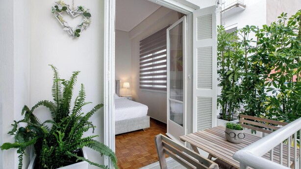 Et lille, lyst værelse med seng, sengebord og et vindue med persienner. Der er en balkon med et træbord og stole, og en potteplante på bordet.