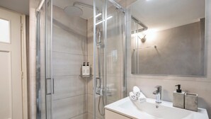 Et moderne badeværelse med en glasafskærmet brusekabine, et hvidt håndvaskarrangement med spejl og toiletartikler.