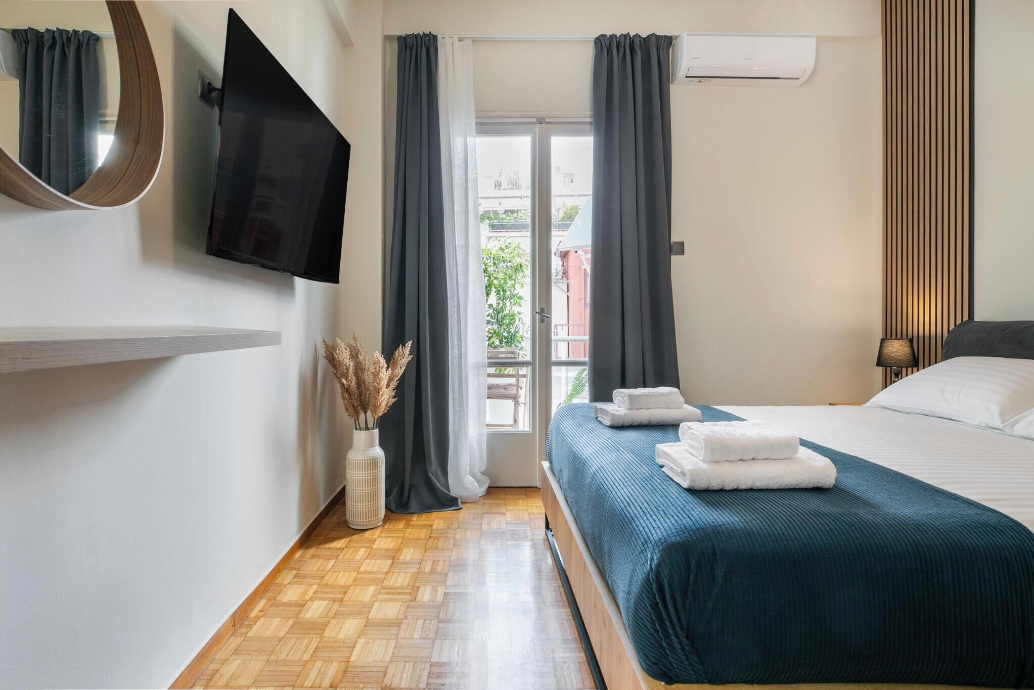 A bedroom with a bed, a flat-screen TV, a window with curtains, a wooden floor, and a wall-mounted air conditioner.