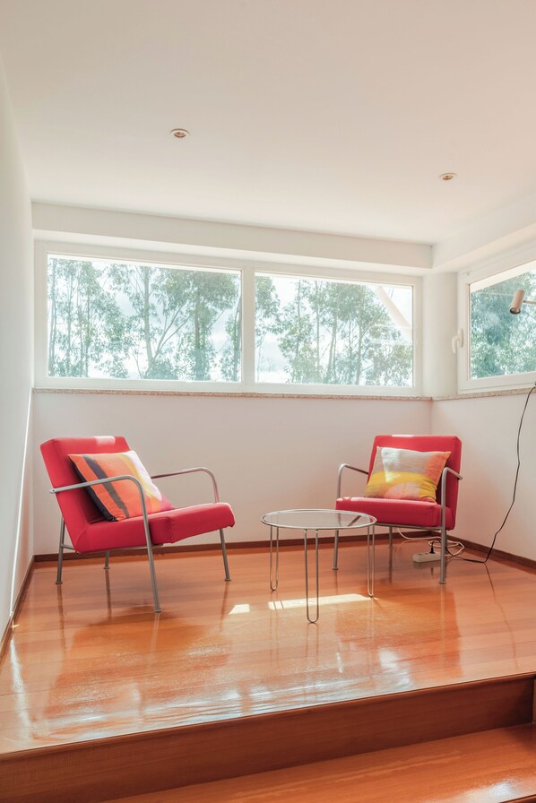 A room with two red chairs, a small table, and large windows with a view of trees.
