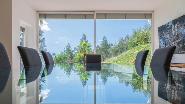 Une pièce moderne avec une table en verre, quatre chaises et une grande fenêtre donnant sur des arbres et une colline.