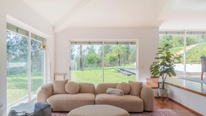 Un salon moderne avec un canapé d’angle beige, un fauteuil poire gris et un pouf rond posé sur un tapis à motifs. De grandes fenêtres offrent une vue sur un jardin luxuriant et un sentier.