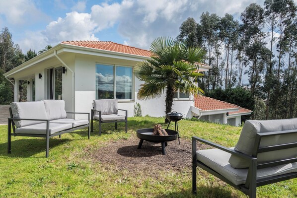 Una casa moderna con un patio que cuenta con dos sofás blancos, un brasero redondo negro y una palmera.