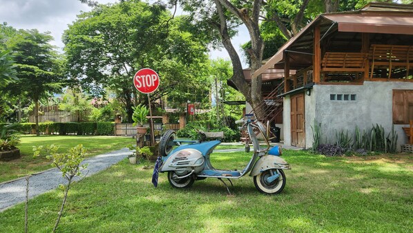 A vintage scooter parked in a lush garden with a stop sign in the background. - Seven 50s (Pai)